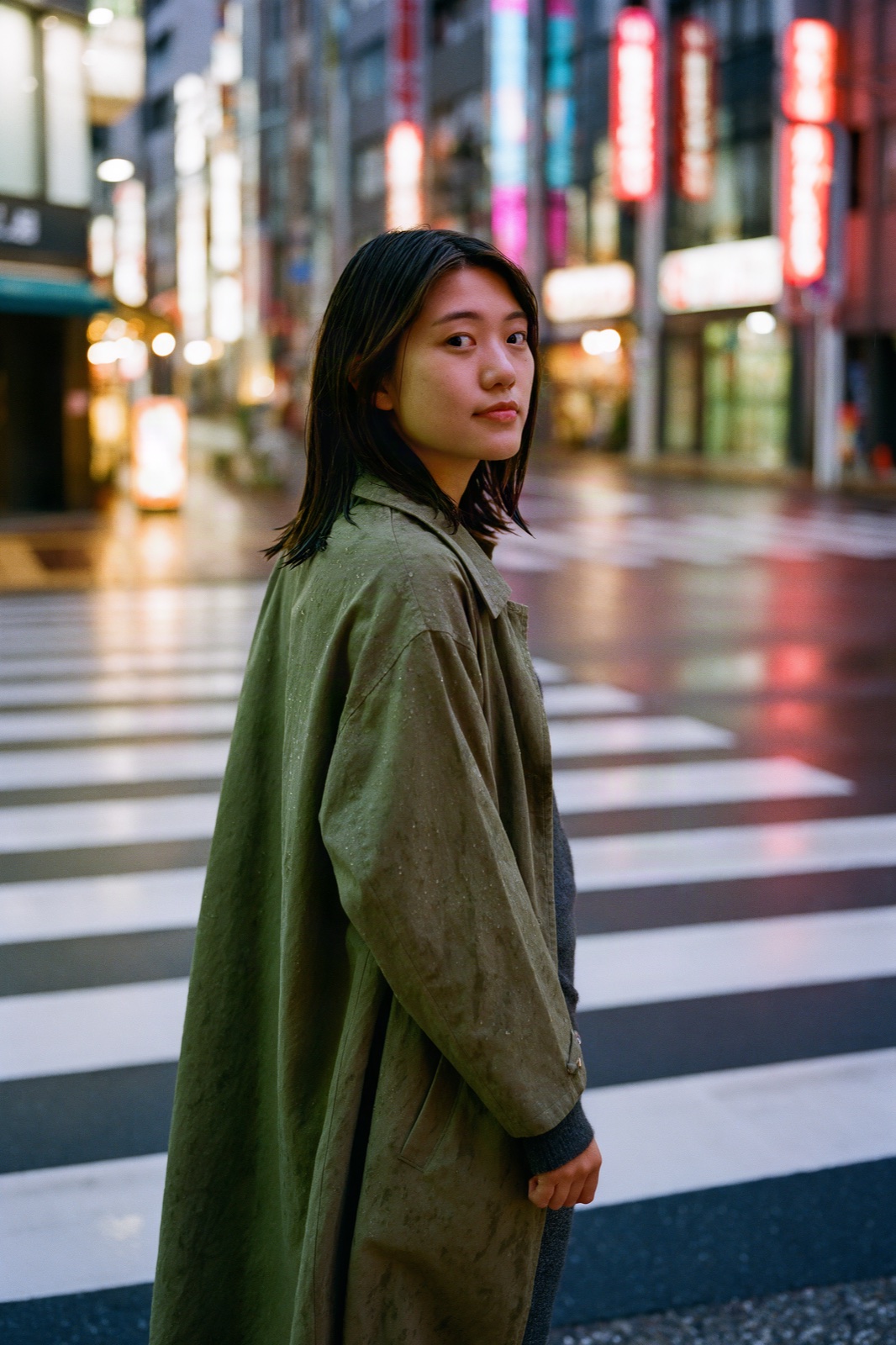 Young woman in an olive trench coat at a Tokyo crosswalk at dusk with neon behind her, Krea 3